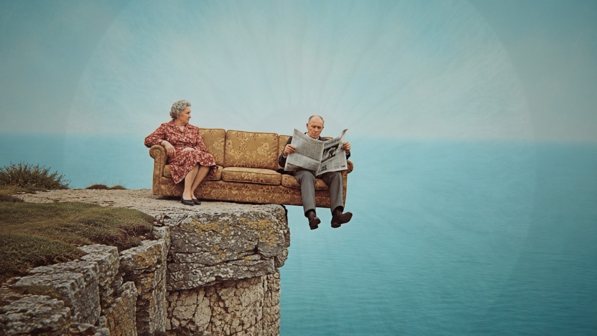 An elderly couple sitting on a floral sofa at the very edge of a cliff, one of them reading a newspaper, the sea filling the rest of the frame.